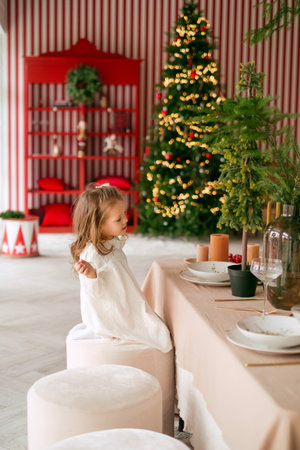 A cute girl is sitting in the living room at a festive table on Christmas Eve. xmas tree with lights in the backgroundの写真素材