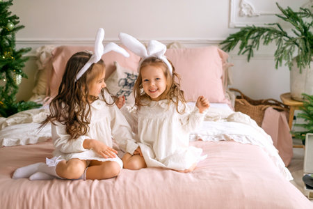 Cute girls with rabbit ears on their heads are sitting on a bed next to a decorated Christmas tree and laughing merrilyの写真素材