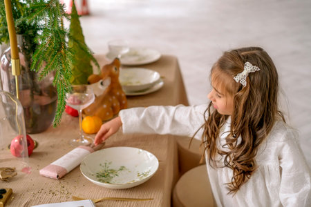 Curly-haired beautiful girl helps to set the festive table for the celebration of Christmas. Daughter lays out the cutlery next to the platesの写真素材