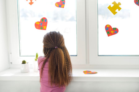 A sad lonely girl looks out the window decorated with heart-shaped postcards. Multicolored puzzle pieces are a symbol of autismの写真素材