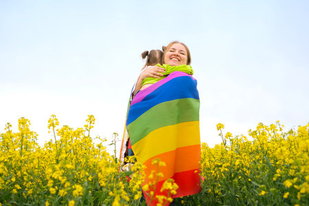 A woman hugs a small child wrapped in a rainbow LGBT flag. Mom and daughter in a field with rapeseed flowersの写真素材