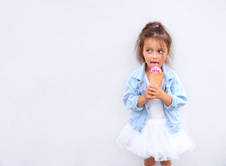 A funny girl stands at the city white wall and licks a pink ball of ice cream looking away. Banner with copy spaceの写真素材