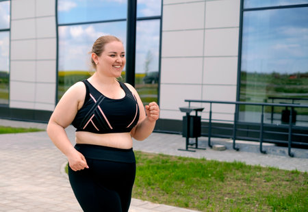 A plump young blonde woman in a sports uniform runs along the sidewalk between tall city buildings. daily morning outdoor sportsの写真素材