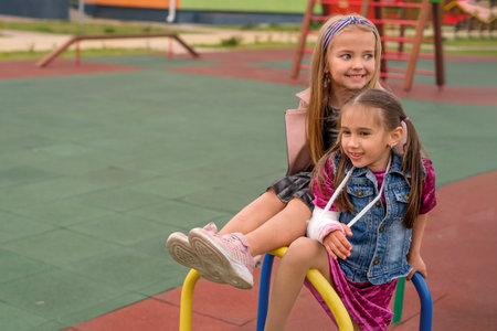 Girl with fractures hand and her friend are sitting on the playground crossbar at the kindergarten.の写真素材