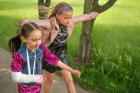 A girl with a fracture and a cast with a bandage on her arm walks carelessly with a friend in the parkの写真素材