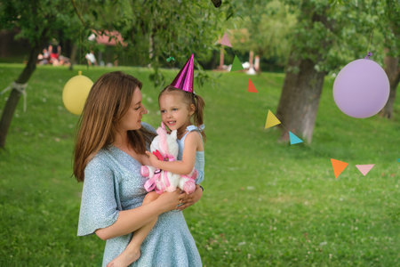 Mom holds a cute beautiful daughter in her arms, giving her a birthday present. A holiday in the park in nature on a warm summer day. The trees are decorated with garlands, flags and balloonsの写真素材