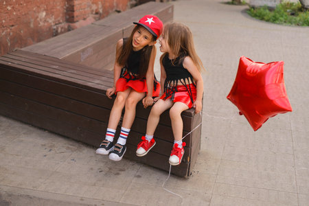 The girl tells interesting rumors and gossip in the ear of a friend sitting on a bench in a city parkの写真素材