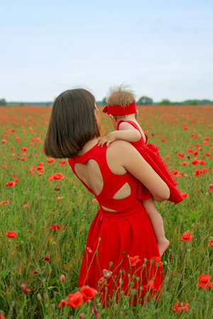 A mother in a red dress with a heart cut out on the back holds her daughter in her arms standing in a meadow with wild poppies growing. mother's dayの写真素材