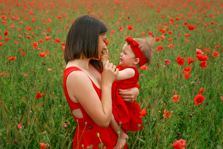 A mother gently kisses her daughter's little hands while standing in a meadow with blooming red poppies. Love, affection, tenderness and care of parents for childrenの写真素材
