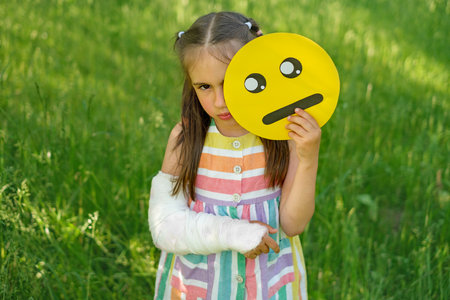 Sad girl with arm injury holds upset emoticon face. Different faces of emojis displaying human emotionsの写真素材