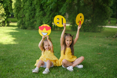 Funny little girls are sitting on the lawn in the park with paper-cut emoji faces. Expression of...