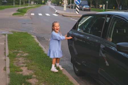 Funny laughing little girl in a blue dress with dreadlocks opens the door to a car parked along a city road on her ownの写真素材
