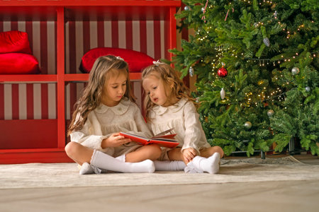 Sisters read New Year's fairy tales sitting on the floor by the Christmas tree. Children look at pictures in a book with storiesの写真素材