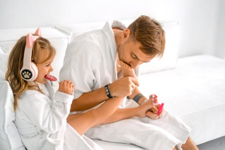 A father lovingly kisses the feet of a cheerful laughing daughter. A girl listens to music with headphones while eating candy.の写真素材