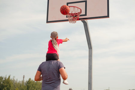 The daughter throws a ball into a basketball basket sitting on her father's shoulders.