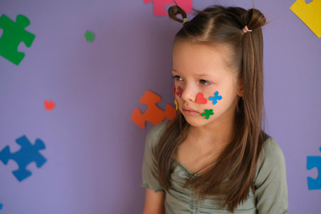 The girl looks away with a sad look. The child's cheeks are decorated with pieces of colored puzzles.の写真素材