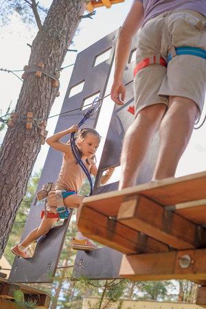 A brave little girl with her father climbs a rope track with suspended steps in an amusement parkの写真素材