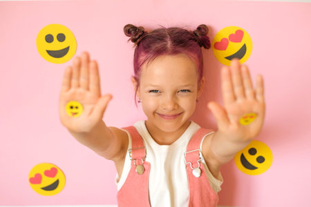 A cute funny girl with pink hair shows emoticons on her palms. A lot of emojis surround the child....