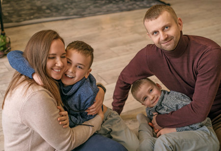 A happy young family with boys is having fun and hugging while sitting on the living room floor....