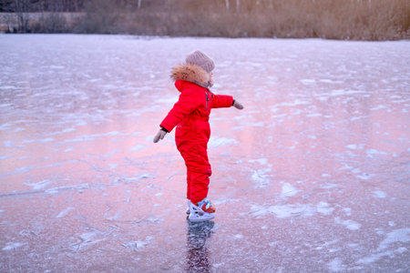 A child in a bright red jumpsuit learns to skate on the ice of a frozen lake at dusk.の写真素材
