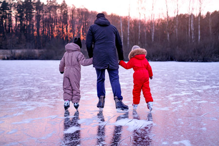 Father holding hands with two children while skating on frozen lake at winter sunset. Family outdoor activity, bonding, care, safety and togetherness in cold season nature landscape.の写真素材