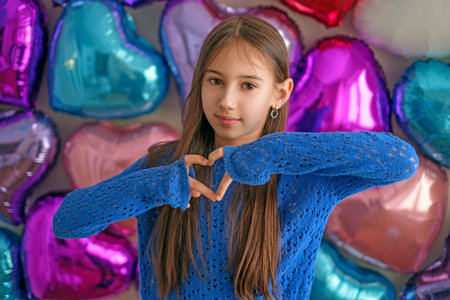 Smiling child making heart shape with hands against colorful heart balloons. Valentine's Day...