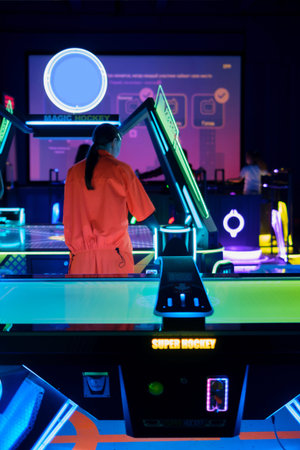 Girl playing air hockey in neon-lit arcade center, viewed from behind. Bright LED lights, colorful...