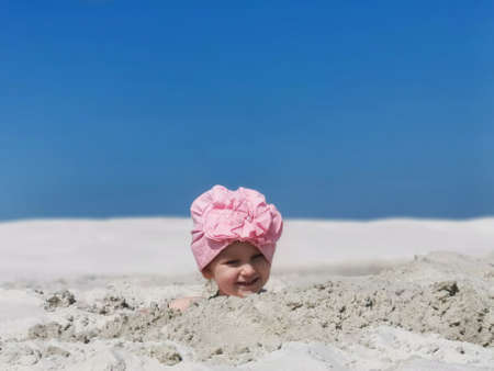 a small child plays in the sand on the beach, buried up to his head, a girl in a scarf smiles while playing, white sand, summer, blue sky, horizontal photo, family vacation, games with childrenの写真素材