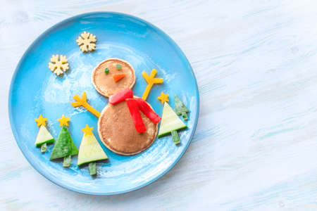 Christmas pancake shaped like a snowman with fresh vegetables blue plate on wooden white table. Christmas fun food for kids, funny breakfast brunch idea xmasの写真素材