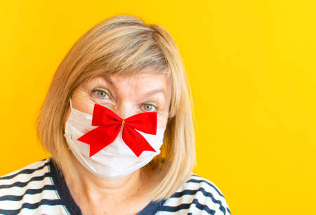 Funny cute senior woman grandmother in protective mask with red christmas bow and new year glasses on isolated yellow background. Quarantine. Stay at home. Helter in place. Coronavirus quarantineの写真素材