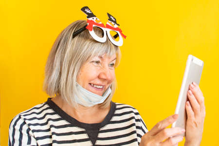 Christmas online congratulations. A woman senior in a medical mask and New Year's glasses with a tablet on a isolated yellow background. Quarantine. Stand at home. Coronavirus quarantine lockdownの写真素材