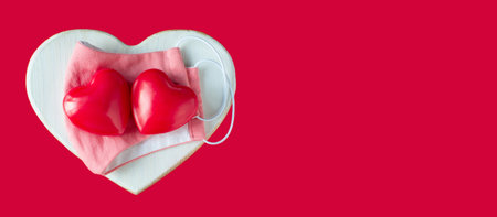 Red hearts with a protective pink mask on a wooden heart on a red background. Valentines day concept care. Banner. Copyspace. Covid-19 Valentine. Coronavirus Valentine. New normal COVID 19.の写真素材