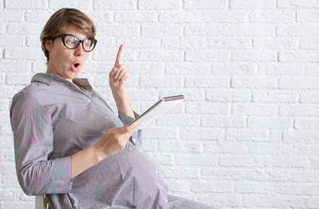 Surprised funny pregnant woman sits, holds a notebook sitting on chair against wall background. Pregnant woman working concept, wishes list, shopping list planning, discussion of childbirth. Copyspaceの写真素材