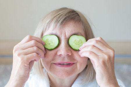 Cheerful senior woman with cucumber slices covering her eyes in front of her eyes in her bedroom. Spa beauty treatments at home, organic cosmetics, body care concept. Natural spa treatment.の写真素材