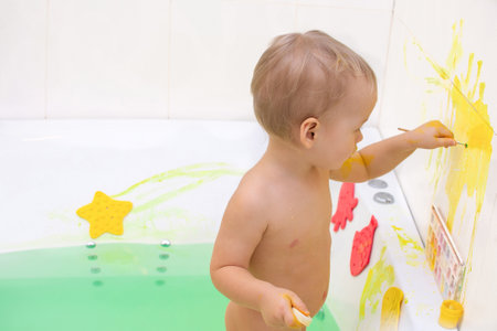 Kid draw with paints in the bath, playing at home. Todler draw with educational toys for creativity. Boy play at home. Bright paint gouache stained face and hands of a child.の写真素材