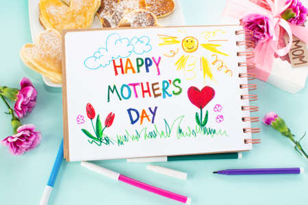 Mothers day breakfast card made by a child. Text Happy Mother day. On a colored background. Pancake in the form of hearts, carnation, gift and a postcard made by the kids for mom.の写真素材