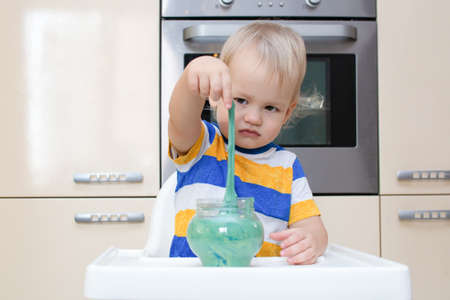 Child play slime at home in the kitchen. Little toddler boy squeeze and stretching green slime sits...