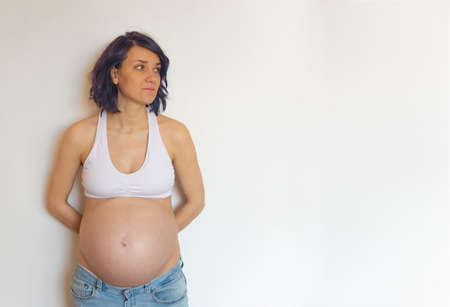 Sad pregnant young girl stands against the background of the wall and looks sadly to the side....