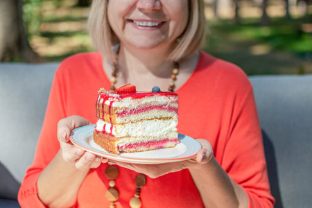 Happy woman eating sweet cake wearing orange clothes in summer park background, holding piece of cake on a plateの写真素材