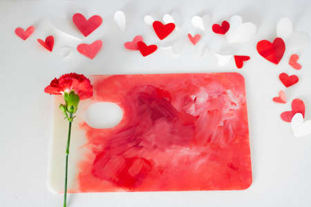 Valentines Day or wedding, invitation, scattered painted small cut out red, pink and white hearts with a red carnation on a colorful paint palette with copy spaceの写真素材