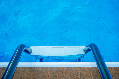 Top step and chrome hand rails of a swimming pool ladder and blue pool water background with copy...