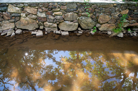 Serene tranquil stone wall by calm water nature background with trees and sky reflected on ancient canal water surface. Shot in natural light with copy space and no people.の写真素材
