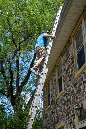 Man on a ladder cleans the gutters on an old Pennsylvania stone home with blue sky and green trees...