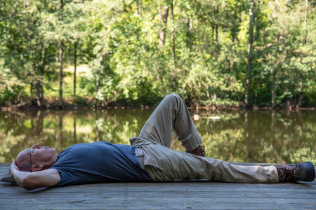 A caucasian senior man rests alone meditating by a calm lake on a wooden dock. Serene nature background. With copy space, shot in natural light.の写真素材