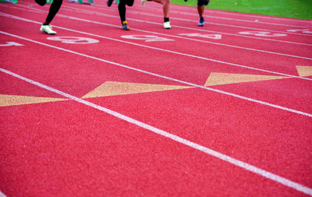 Bright red textrued athletic track with crisp white lane lines and markings with white numbers. Four people race toward the camera. With copy space and selective focus.の写真素材