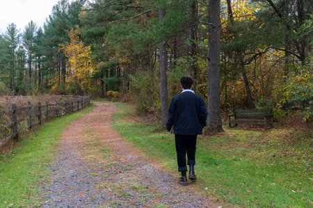 One young man wears black and walks away alone down an autumn country road with gravel, grass, a...