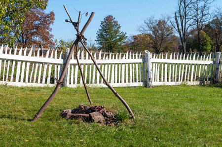 A white picket fence in the background and a backyard with a cook fire with tripod for hanging a...