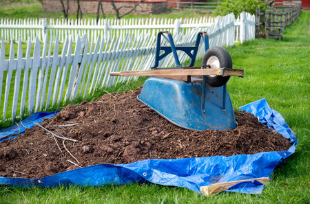 Beautiful rustic grass and white picket fence with a pile of garden soil on an outstretched blue tarp and an upturned weathered blue metal wheelbarrow on top.の写真素材
