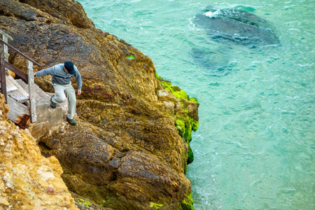 Man climbs onto cliff rocks at base of hiking trail steps by ocean Salema Beach Algarve Portugalの写真素材