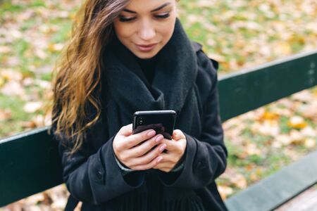 Beautiful girl sitting on a bench, woman with your phone writes a message on social networks, sunny autumn day in the park, relaxing, after work business woman.の写真素材